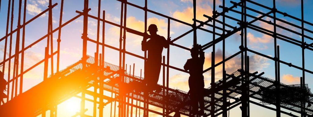 Operai al lavoro su un ponteggio in un cantiere edile al tramonto, con strutture metalliche e dispositivi di sicurezza.